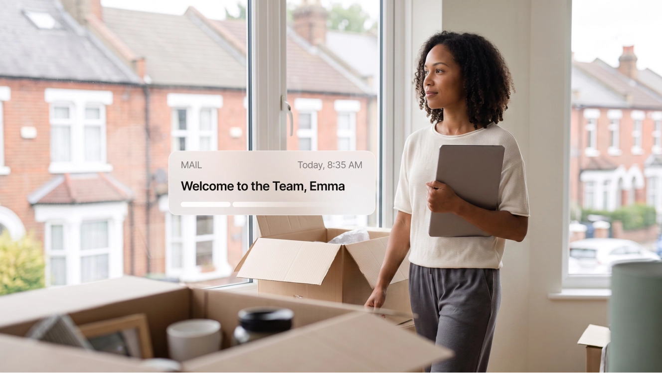 A new hire standing among moving boxes by a window overlooking UK terraced houses, with a welcome email notification